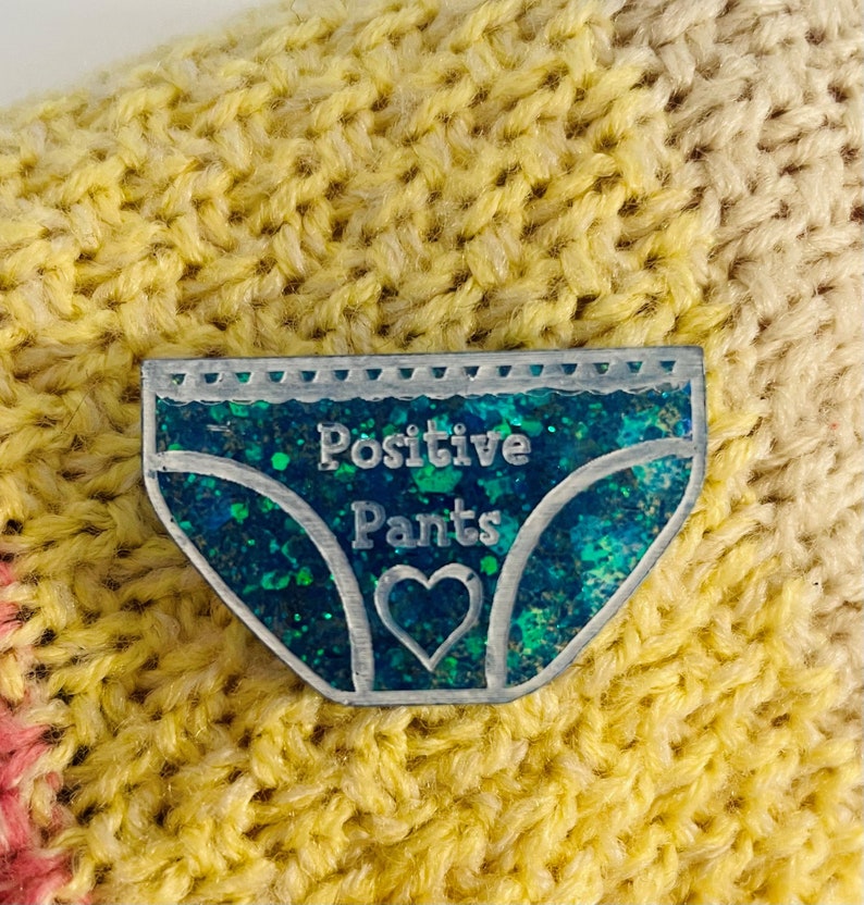 Pu&ograve; includere: Uno spillo di glitter blu e verde a forma di biancheria intima con il testo "Positive Pants" e un cuore al centro.