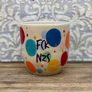 May include: A cream-colored ceramic mug featuring a hand-painted pattern of multicolored polka dots in shades of blue, red, orange, and yellow. The text F(K NZS is written in black, bold, hand-lettered font across the center of the mug surface.