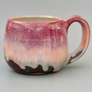May include: A handmade ceramic mug with a rounded body and a large handle. The mug features a gradient of colors, transitioning from deep red at the top to light pink and white, with a dark brown base and green accents. The mug is designed for hot beverages.