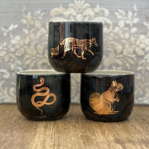 Peut inclure: Trois tasses cylindriques en céramique avec une finition noire brillante. Chaque tasse présente une illustration métallique de couleur cuivre : un renard en marche, un serpent enroulé et un écureuil assis. Les tasses ont une texture lisse.
