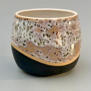 Könnte beinhalten: Ein handgefertigter Keramikbecher mit einer einzigartigen, gesprenkelten Glasur. Der Becher hat einen schwarzen Boden, ein beiges Band und einen gesprenkelten weißen und beigen oberen Bereich. Der Innenraum ist cremeweiß. Ideal für Kaffee oder Tee.
