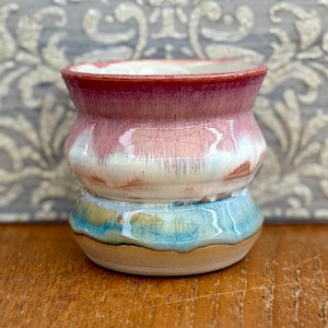 May include: A small ceramic cup with a rounded, bulbous shape. The glaze features a gradient transition from a deep reddish-pink at the rim, blending into a creamy white middle section, and finishing with a light blue and tan base.