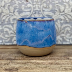 May include: A rounded ceramic cup featuring a glossy blue glaze with subtle white wavy patterns. The upper portion is a vibrant blue, while the lower section remains an unglazed, matte beige clay, creating a distinct contrast in texture.