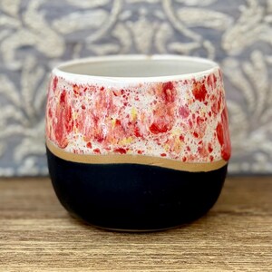 May include: A ceramic cup featuring a rounded shape with a two-tone design. The upper portion displays a speckled pattern of vibrant red, orange, and white glaze, while the lower portion is finished in a solid matte black glaze.