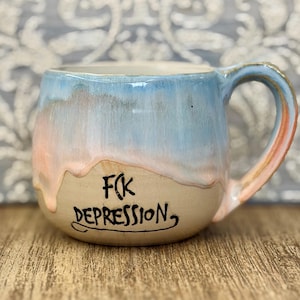 Könnte beinhalten: Ein Keramikbecher mit abgerundeter Form, einer beigen Basis und einer hellblauen sowie pfirsichfarbenen Glasur, die an den Seiten herabläuft. Der Text F(K DEPRESSION ist in schwarzer, handgeschriebener Schrift auf der Vorderseite zu sehen.