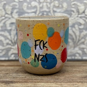 Könnte beinhalten: Ein gesprenkelter beiger Keramikbecher mit einem Muster aus bunten, gemalten Kreisen in Gelb, Orange, Rot, Türkis und Blau. Der Text FCK NZS ist in schwarzen Buchstaben auf der Vorderseite des Bechers aufgedruckt, der eine glatte, abgerundete Form hat.