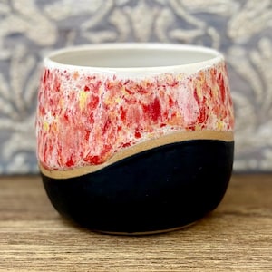 Könnte beinhalten: Ein Keramikbecher mit zweifarbigem Design, bestehend aus einer glatten, mattschwarzen Basis und einem texturierten oberen Teil. Der obere Bereich zeigt eine lebendige, gesprenkelte Glasur in leuchtendem Rot, Weiß und Gelb, getrennt durch eine Kurve.