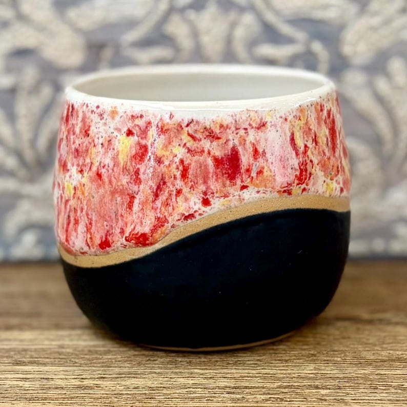 May include: A ceramic cup featuring a two-tone design with a smooth matte black base and a textured upper section. The top portion displays a vibrant, mottled glaze in shades of bright red, white, and subtle yellow, separated by a curved line.