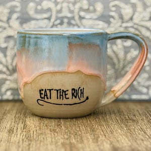 Könnte beinhalten: Ein Keramikbecher mit abgerundeter Form, der einen hellblauen und zartrosa glasierten oberen Bereich aufweist, der über eine naturfarbene beige Basis läuft. Der Text "EAT THE RICH" ist in schwarzer Tinte auf der Vorderseite aufgedruckt.