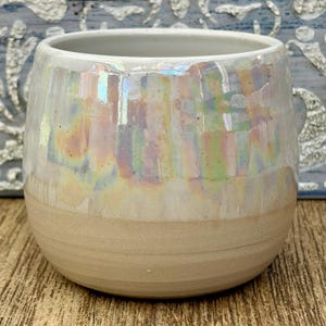 Puede incluir: Una taza de cerámica redondeada con una base beige lisa sin esmaltar y una sección superior brillante con un acabado iridiscente y nacarado que refleja colores pastel suaves, incluyendo rosa, azul y amarillo, sobre su superficie curva.