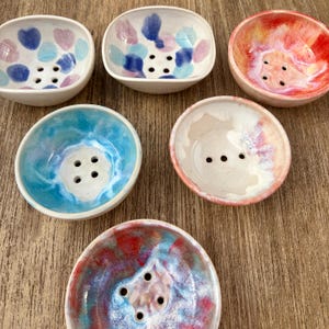 Może przedstawiać: Kolekcja ręcznie robionych ceramicznych mydelnic w różnych kształtach i kolorach. Naczynia mają różne wzory, w tym serca i abstrakcyjne wzory. Każde naczynie ma otwory drenażowe i jest glazurowane żywymi odcieniami niebieskiego, różowego i czerwonego.