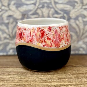 May include: A ceramic cup with a rounded shape featuring a two-tone design. The upper portion has a speckled, textured glaze in shades of red, orange, and white, while the lower portion is a solid, matte black finish with a smooth, curved transition.