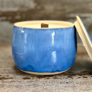 Könnte beinhalten: Eine blaue Keramikkerze in einem runden Behälter mit einem Holzdocht. Die Kerze hat eine glänzende Oberfläche und einen hellfarbigen Rand. Der Deckel ist teilweise sichtbar, und der Hintergrund ist neutral.