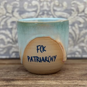 Könnte beinhalten: Ein Keramikbecher mit abgerundeter Form, einer natürlichen beigen Basis und einer hellblauen Glasur, die vom Rand herabläuft. Der Text FCK PATRIARCHY ist in dunkelblauen Buchstaben auf der Vorderseite des Bechers im beigen Bereich zentriert.