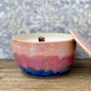 May include: A round ceramic candle vessel featuring a gradient glaze that transitions from a soft peach at the top to a vibrant pink in the middle and a deep blue at the base. The candle has a single wooden wick and a matching ceramic lid.