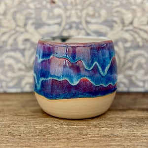 Könnte beinhalten: Ein abgerundeter Keramikbecher mit einer glänzenden Glasur, die in Schichten aus tiefem Blau, Violett und Türkis verläuft. Der obere Teil zeigt ein lebhaftes, fließendes Muster dieser Farben, während der untere Boden glatt und unglasiert beige ist.