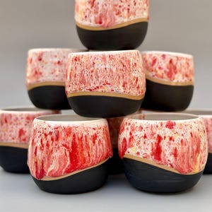 Può includere: Una pila di tazze in ceramica fatte a mano. Le tazze hanno una base nera, una fascia color oro e una sezione superiore con una glassa maculata rossa e bianca. Le tazze sono disposte a piramide, mostrando il loro design unico.