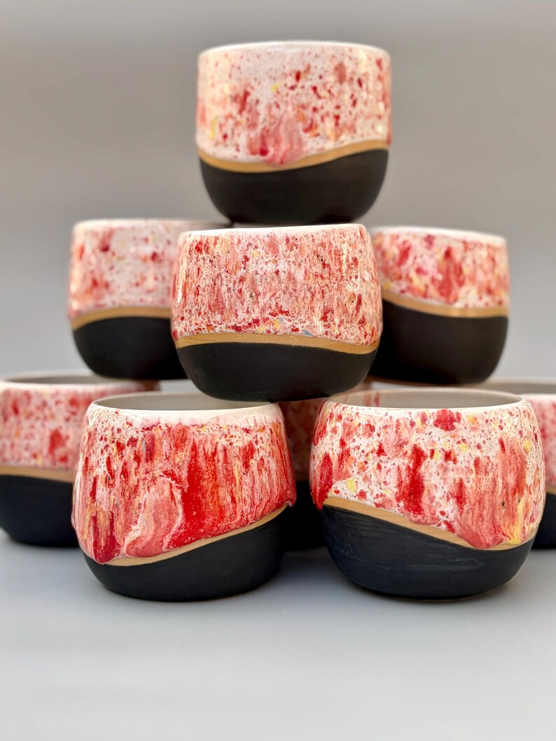 May include: A stack of seven ceramic cups featuring a two-tone design. The upper portion of each cup has a speckled, marbled pattern in shades of red, white, and yellow, while the lower portion is a solid matte black with a curved, wavy border.