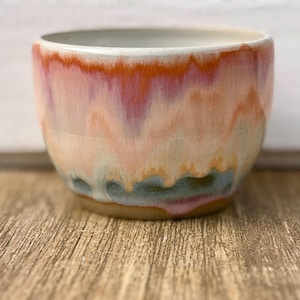Puede incluir: Un cuenco de cerámica hecho a mano con un esmalte único. El cuenco presenta una mezcla de colores, incluyendo melocotón, naranja y azul, creando un efecto acuarela. El cuenco es redondo y tiene un acabado liso y brillante. Perfecto para servir comida o como pieza decorativa.