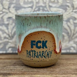 Könnte beinhalten: Ein Keramikbecher mit gesprenkelter beiger Basis und hellgrüner Tropfglasur am oberen Rand. Der Text FCK PATRIARCHY ist in dunkelblauen Buchstaben auf der Vorderseite des Bechers aufgedruckt. Der Becher hat eine abgerundete Form und eine glatte Glasur.