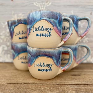 May include: A collection of ceramic mugs featuring a cream-colored base and a blue and pink drip glaze at the top. Each mug has a handle and the text "Lieblingsmensch" printed in dark blue script on the front center of the smooth surface.