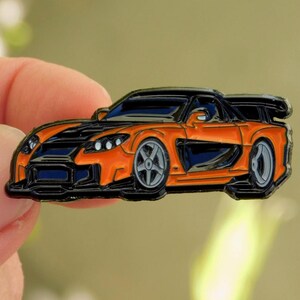 May include: Orange and black sports car enamel pin. The pin features a detailed design of a sleek, low-profile car with a spoiler. The car has silver wheels and black accents. The pin is outlined in black.