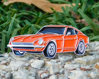 Könnte beinhalten: Orangener Emaille-Pin einer klassischen Sportwagen mit silbernen Zierleisten und schwarzen Details. Das Auto hat ein schlankes, aerodynamisches Design mit detaillierten Rädern. Der Pin befindet sich vor einem Hintergrund aus Steinen und grünem Gras.