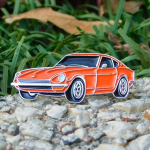 Puede incluir: Pin de esmalte naranja de un coche deportivo clásico con detalles plateados y negros. El coche tiene un diseño elegante y aerodinámico con ruedas detalladas. El pin está sobre un fondo de rocas y hierba verde.