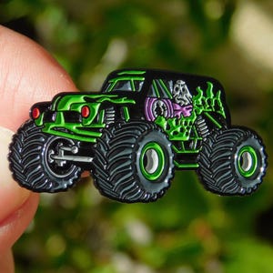 May include: A green and black monster truck pin with a skull graphic. The pin features large black tires with green accents, red headlights, and a detailed design. The truck has a black body with green highlights.