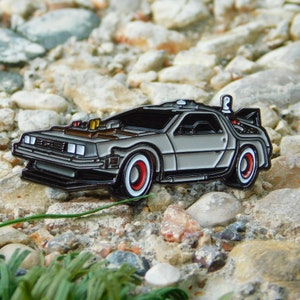 May include: A silver and black enamel pin of the DeLorean time machine from the film Back to the Future. The pin has red and white wheels and a black outline.