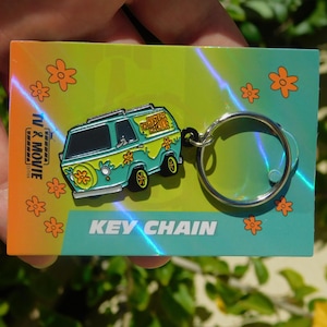 May include: A colorful keychain featuring the Mystery Machine van from the TV show. The van is teal and yellow with a flower design. The keychain is attached to a card that says "KEY CHAIN" and has orange flowers.