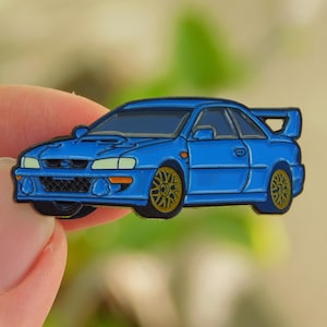 May include: A blue car pin with gold wheels and a rear spoiler. The pin is a stylized representation of a sports car, with detailed headlights and a dark blue outline. The car is held between fingers against a blurred background.