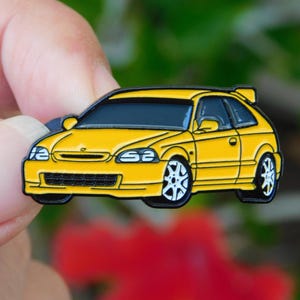 Puede incluir: Pin de esmalte de coche amarillo con ribete negro y ruedas blancas. El pin presenta un diseño detallado de un cupé con alerón. El coche tiene ventanas negras y una parrilla delantera negra. El pin lo sostiene una persona.
