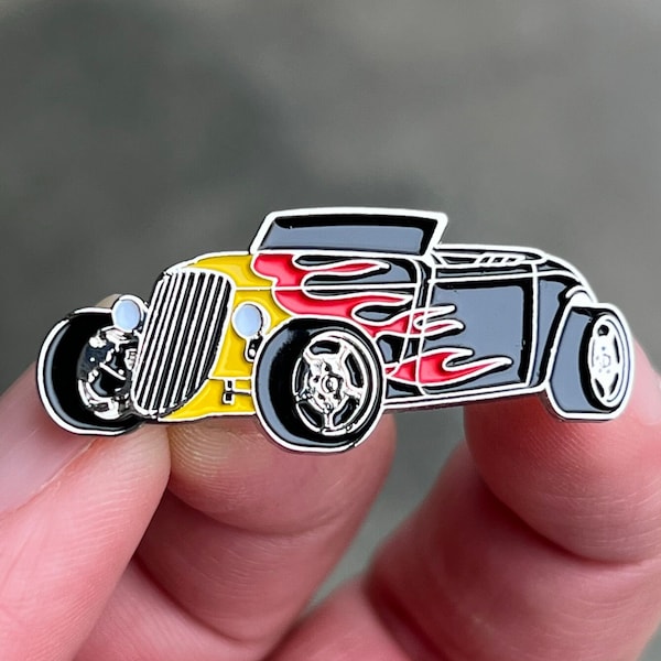 May include: A yellow and black enamel pin of a vintage hot rod with red flames. The car has a black grille and chrome wheels.