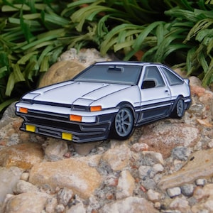 Puede incluir: Pin de esmalte blanco de un coche deportivo japonés clásico con detalles negros y faros amarillos.