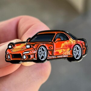 May include: Orange and yellow enamel pin of a red Mazda RX-7 with black accents and a J-Spec Toyo Tires logo. The car has a black interior and black wheels with white spokes. The pin has a black outline.