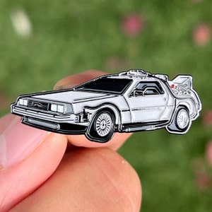 Pode incluir: Um broche de esmalte branco de uma máquina do tempo DeLorean do filme De Volta para o Futuro. O broche é detalhado com detalhes pretos e tem uma luz vermelha na parte de trás.