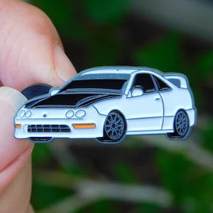 May include: A white and black car-shaped enamel pin. The pin features a white body with a black hood and dark gray wheels. The car has orange accents and is held by a person's fingers.