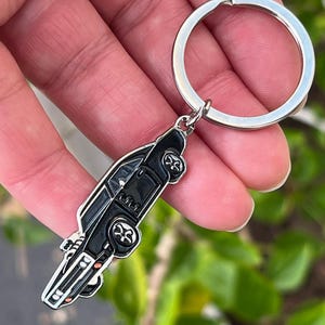 May include: A black car keychain with a silver key ring. The car has detailed wheels and a sleek design. The keychain is held up against a background of green foliage.