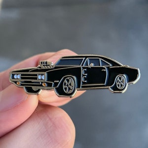 May include: A black enamel pin of a classic muscle car, a Dodge Charger, with a supercharger and black wheels. The pin is a detailed representation of the car.