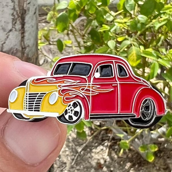 May include: An enamel pin depicting a vintage car, a 1940s Ford Coupe, in red and yellow with flames on the side.