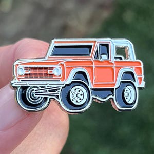 Peut inclure: Épinglette en émail orange représentant un Ford Bronco vintage avec une calandre noire et des finitions argentées.