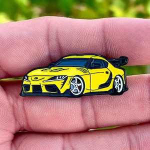 May include: A yellow enamel pin of a Toyota Supra sports car with black accents and a black spoiler. The car has black wheels with red accents.