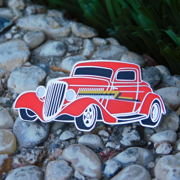 May include: A red vintage car pin with chrome accents and a black grille. The car has a yellow and black stripe on the side. 