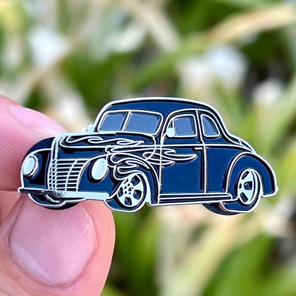 May include: Black enamel pin of a vintage car with flames painted on the side. The car is a 1940s Ford Coupe.