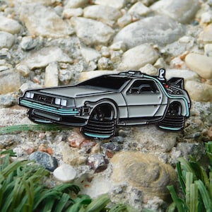 May include: A silver and black enamel pin of a DeLorean car, featuring black wheels and light blue accents. The pin is set against a backdrop of rocks and small stones, giving it a rugged appearance.