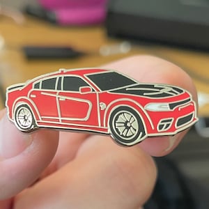 Pode incluir: Um broche de esmalte vermelho e prata de um carro Dodge Charger. O carro tem um capô escurecido e rodas pretas.