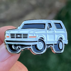 Puede incluir: Un pin de esmalte plateado de un Ford Bronco blanco. El pin está detallado con acentos negros y una parrilla negra.