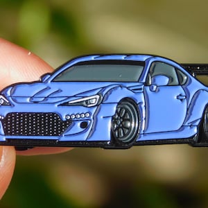 Op de afbeelding: Een blauwe sportwagen emaille pin met zwarte accenten. De pin heeft een gedetailleerd ontwerp van een aangepaste auto met een spoiler, zwarte wielen en een zwarte grille. De auto is een levendige tint blauw.