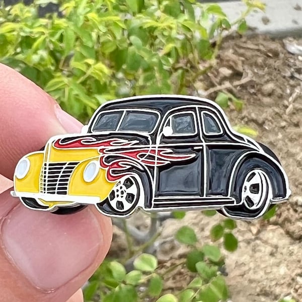 May include: A black and yellow enamel pin of a vintage car with red flames on the side. The car has a black body, yellow wings, and chrome wheels.